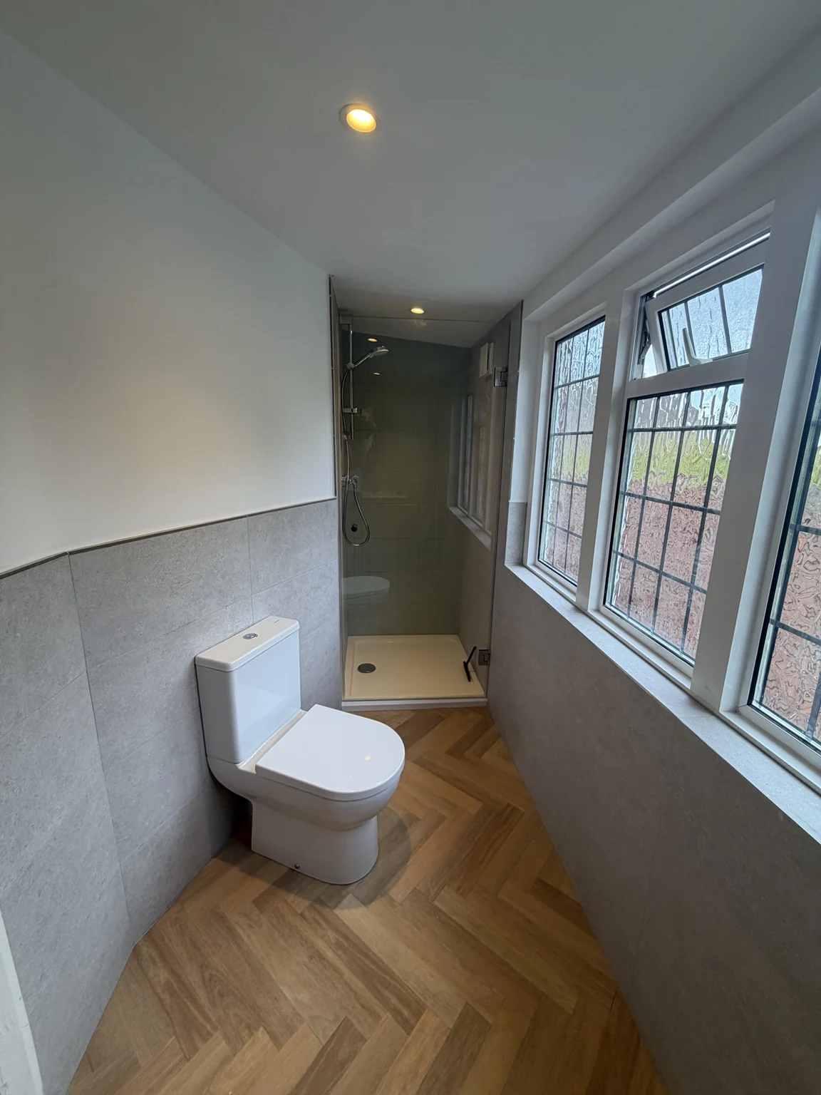 Finished ensuite showing the WC, glass shower enclosure, grey wall tiles and herringbone-pattern flooring.